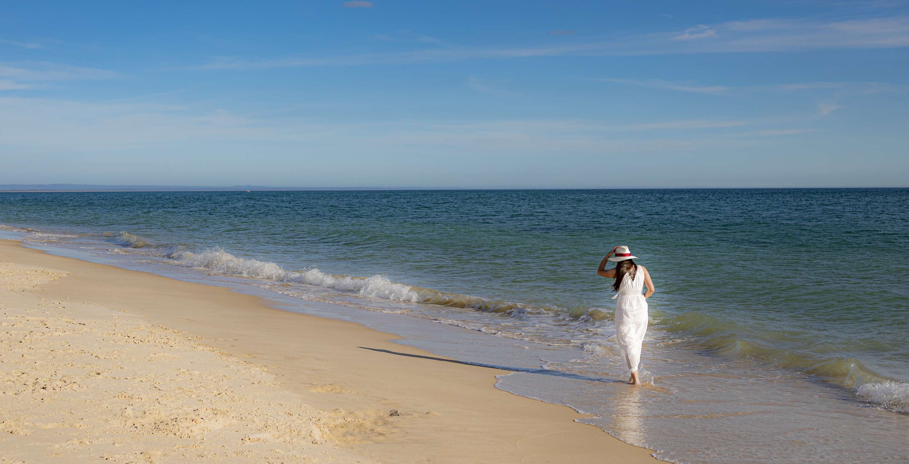 Woman in white dress and hat walks by the sea near Pestana Tróia Eco-Resort, with direct beach access