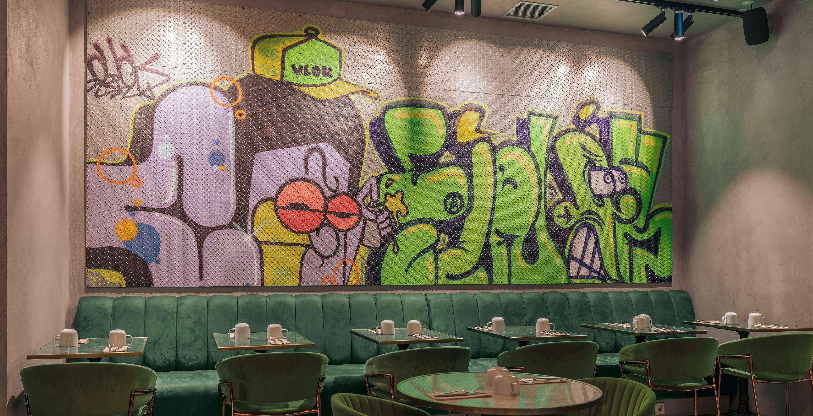 Restaurant with green tables, chairs and sofas, with white china glasses and a colorful wall with graffiti