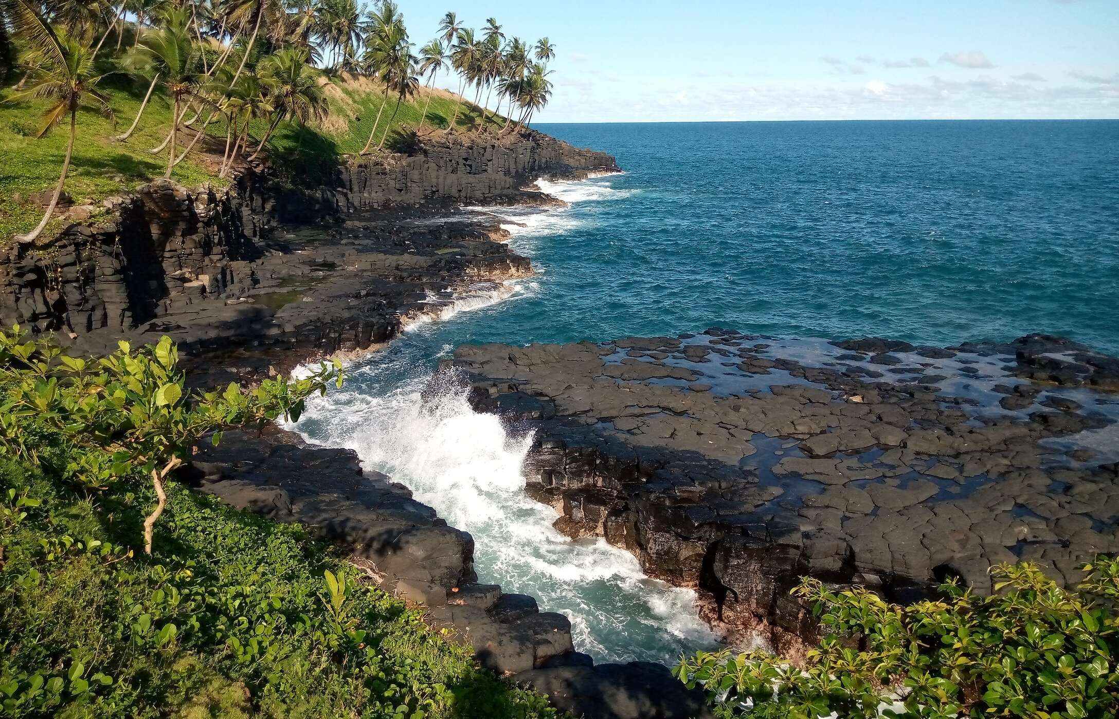 Vista de la Boca del Infierno en Santo Tomé y Príncipe, con mar agitado, rocas negras y vegetación alrededor.