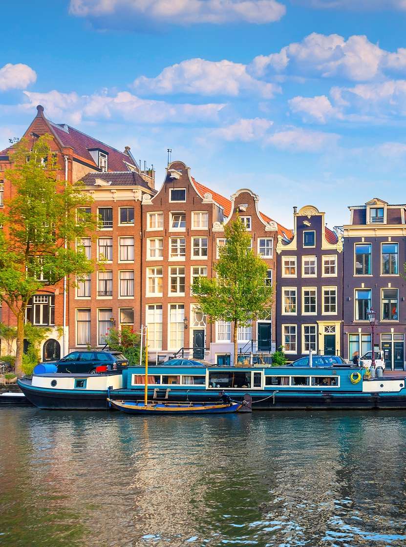 Canal de Ámsterdam con gente caminando por la orilla, barcos y edificios típicos bajo un cielo azul