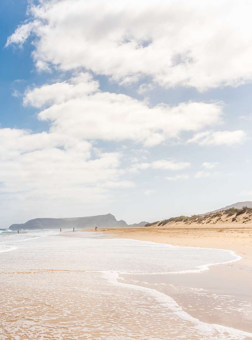 Praia extensa em Porto Santo com areia clara, ondas calmas, e um céu azul com alguma nuvens