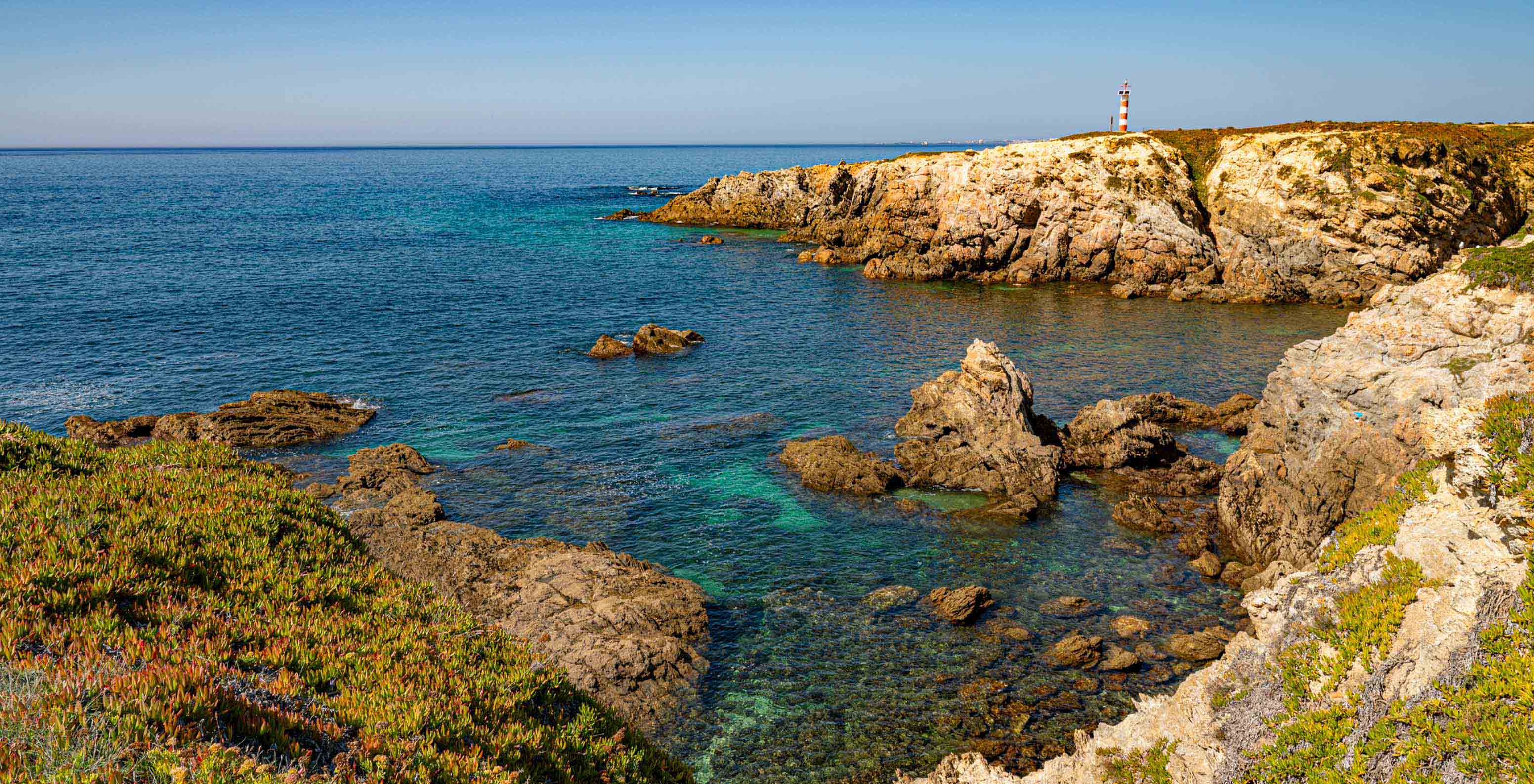 Vista costeira rochosa com águas azul-turquesa e um farol ao fundo perto do Pestana Porto Covo Village