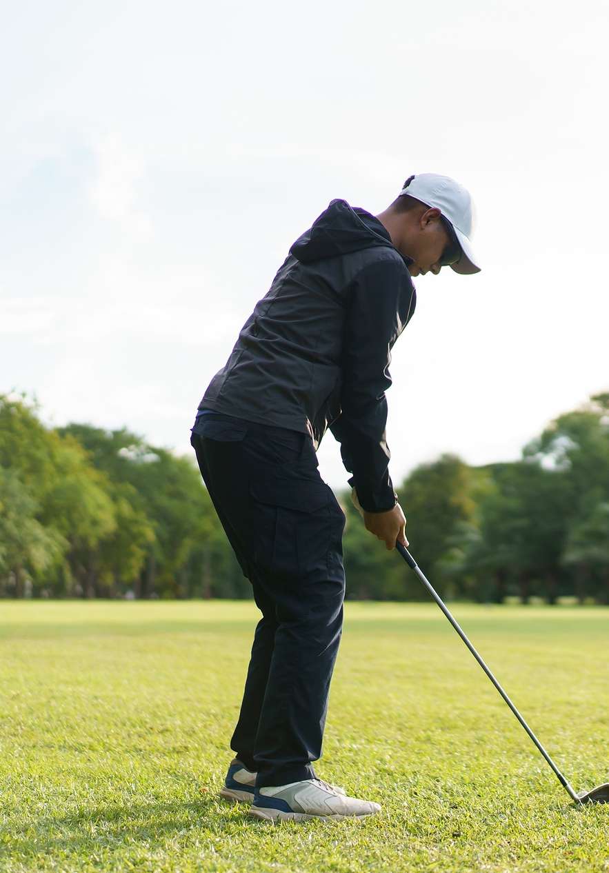 Homem de chapéu branco e roupas desportivas a jogar golfe num campo muito verde com árvores desfocadas ao longe