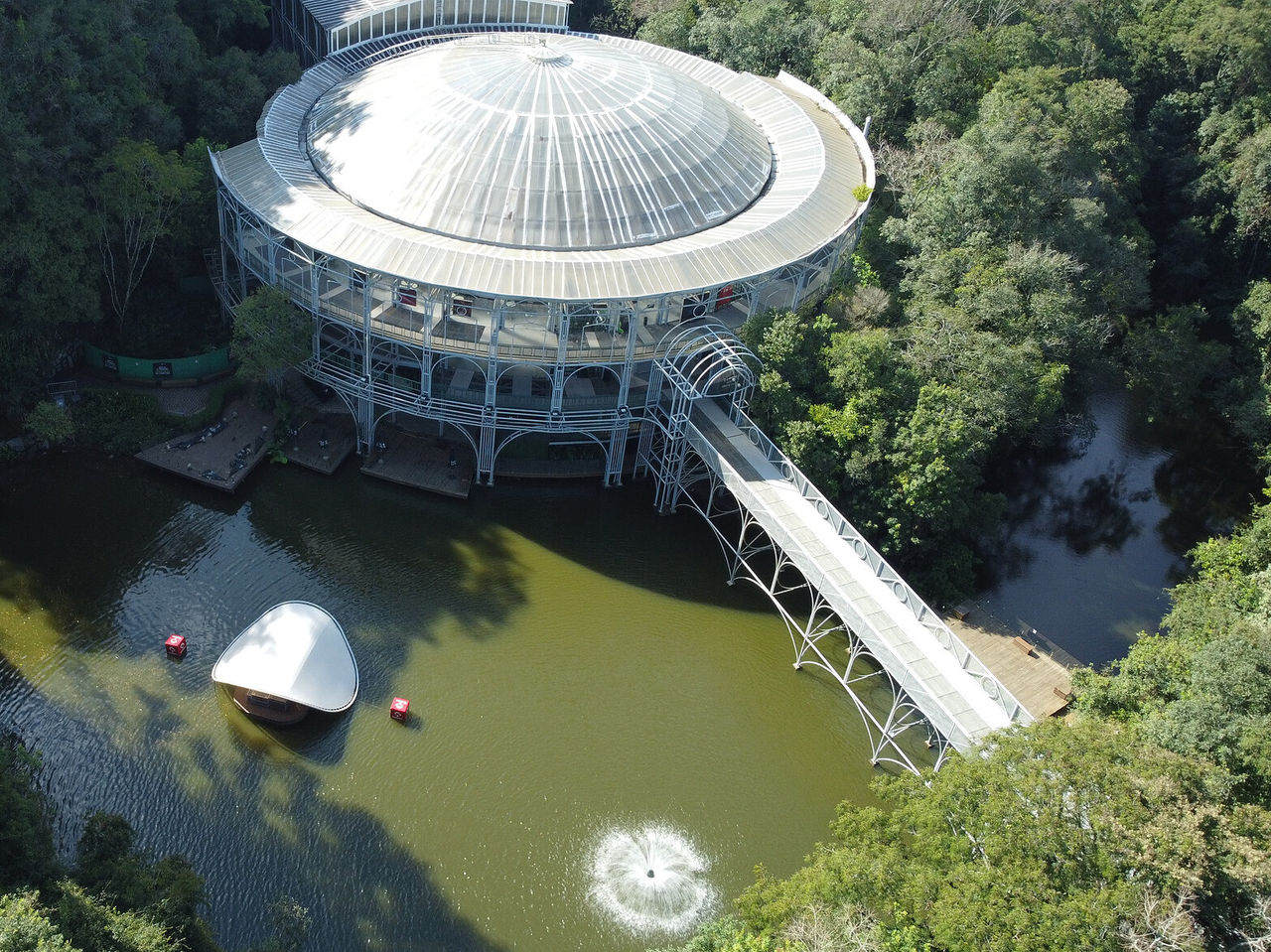 Luchtfoto van de Ópera de Arame, met zijn iconische ronde structuur, omgeven door natuur, met een brug en een meer