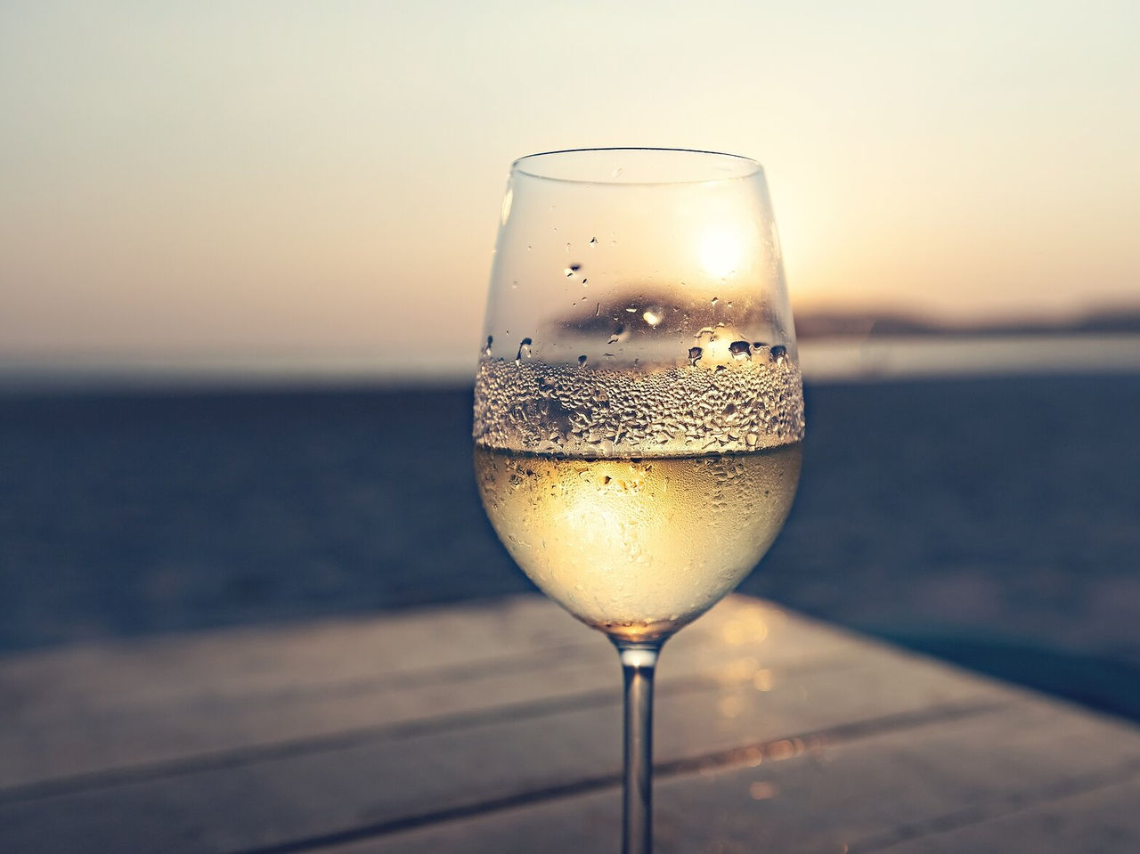 Glass of white wine with blurred sunset reflections in the background, on a wooden surface